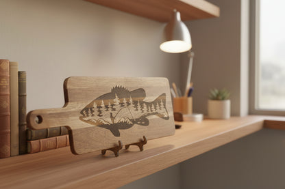 A rectangular wooden cutting board with a handle, featuring a laser-engraved bass fish design with a forest and fishing rod motif. The board is displayed on a small stand against a gray textured wall.