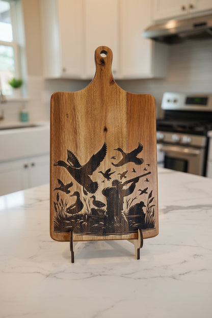 A rectangular wooden cutting board with a handle, featuring a dark engraved design of birds and reeds on one side, photographed indoors on a gray floor.